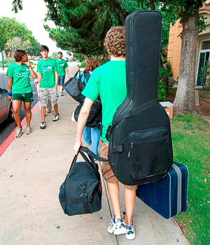 Student with a large acoustic guitar case worn like a backpack and luggage in each hand walking to a dorm.