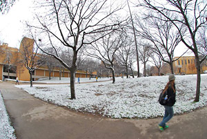 Student walking on the sidewalk with snow on the surrounding grass.