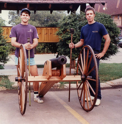Rick and Alex stand ready behind Boomer, a muzzle loading cannon with wooden frame and spoked wheels.