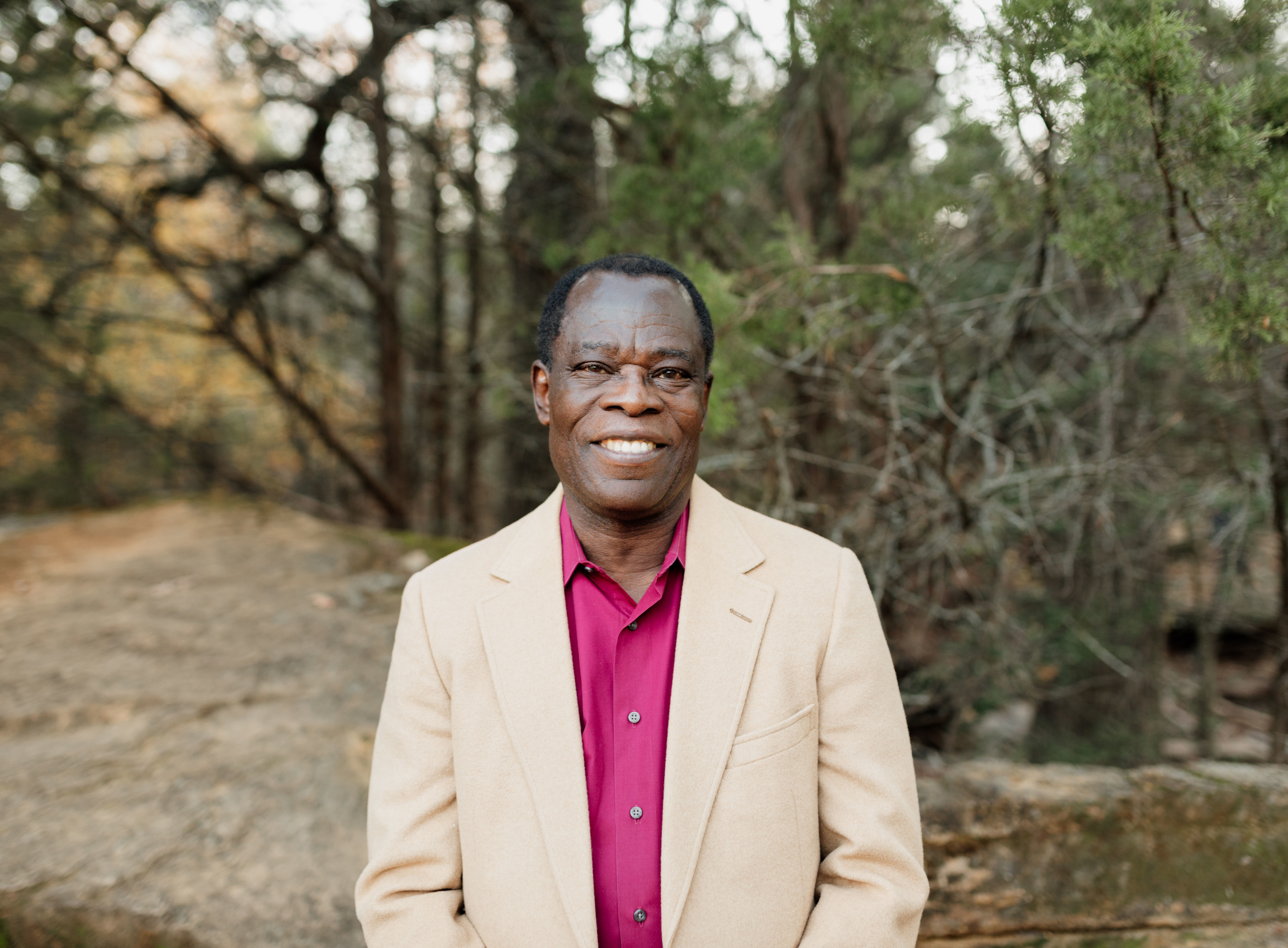 Joseph Oppong stands in a pink shirt and beige jacket in front of trees.