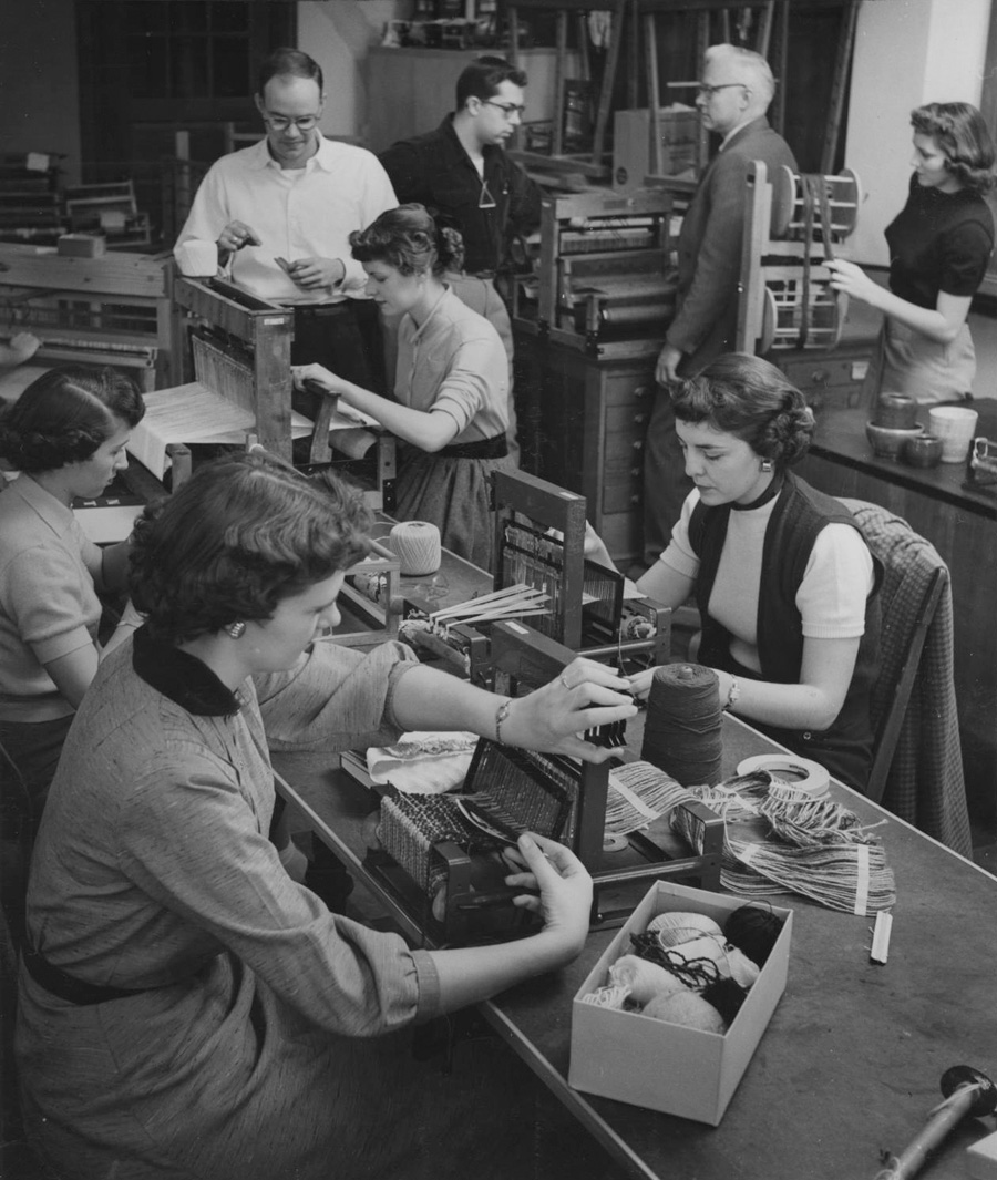 Four women are seated at their workstations and working at their looms