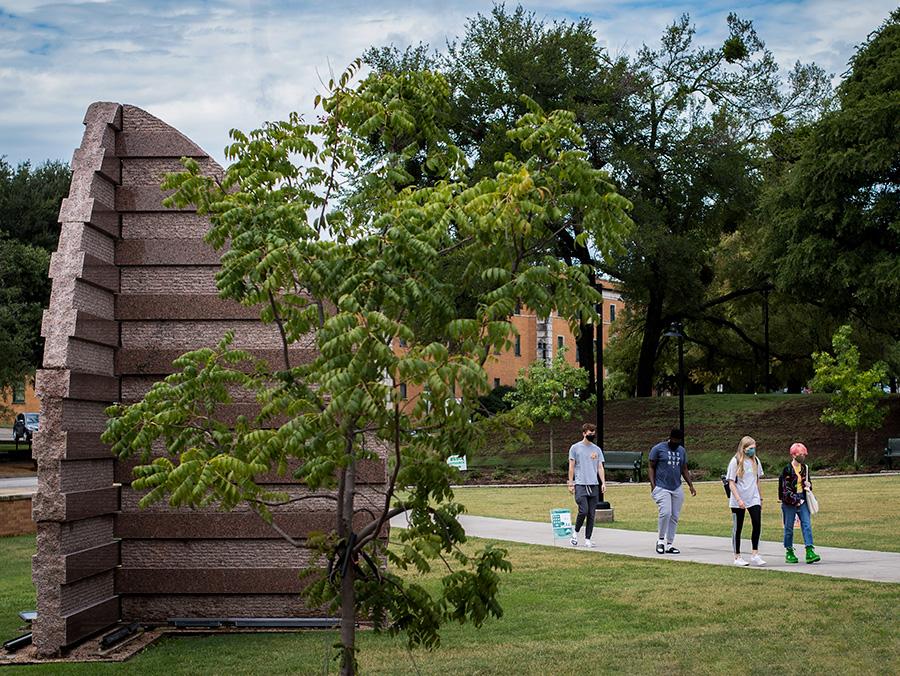 Picture of students walking outside of UNT campus with the sculpture "Shield" in the foreground and trees on both sides.