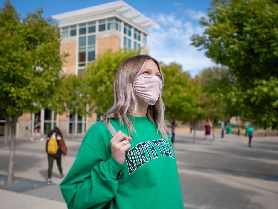 Blond female student in green North Texas sweatshirt walks in front of the University Union holding a bag.