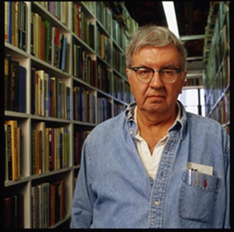 Larry McMurtry wears a blue shirt and stands in front of rows of bookshelves.